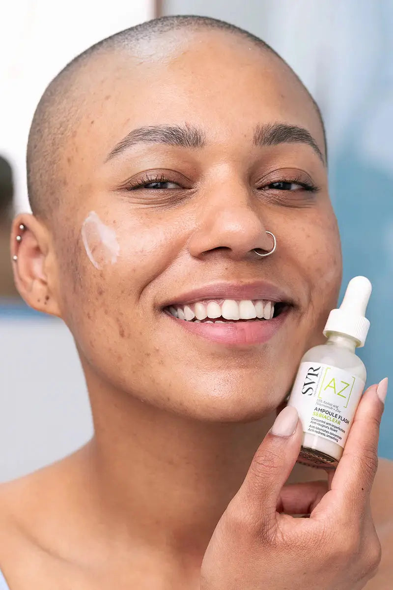 A close-up of a smiling person with a shaved head holding a small skincare dropper bottle next to a white cream smear on their cheek. The bottle label shows the brand STRAZ.