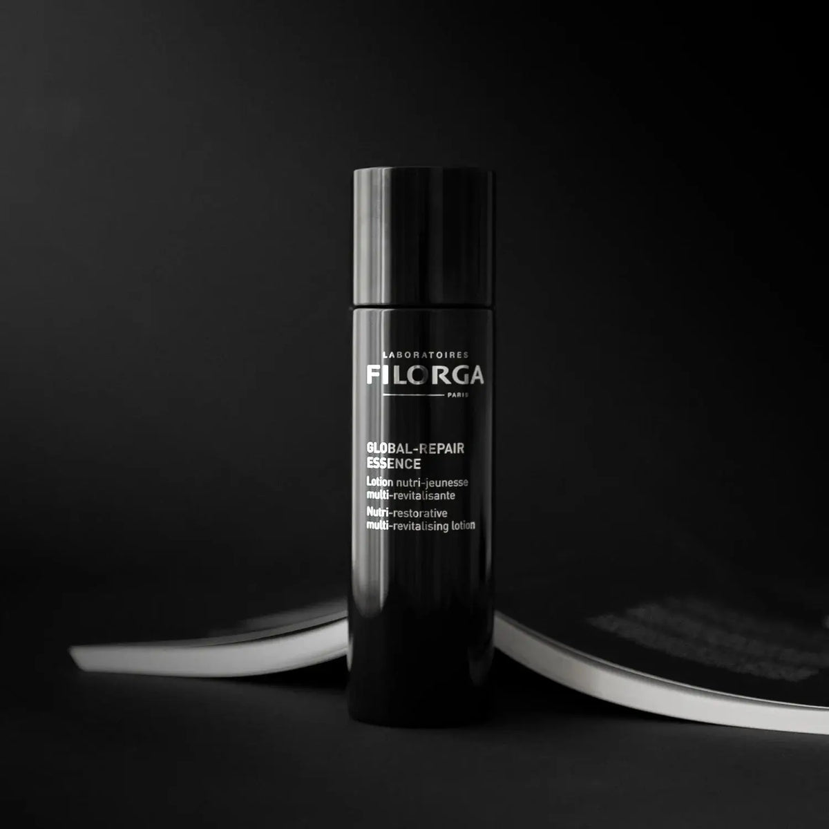 A black cylindrical bottle of Fila RGA Global J-Ehair Essence stands upright on a dark surface with a blurred magazine in the background.