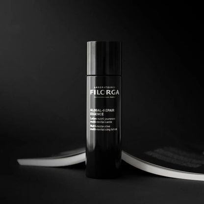 A black cylindrical bottle of Fila RGA Global J-Ehair Essence stands upright on a dark surface with a blurred magazine in the background.
