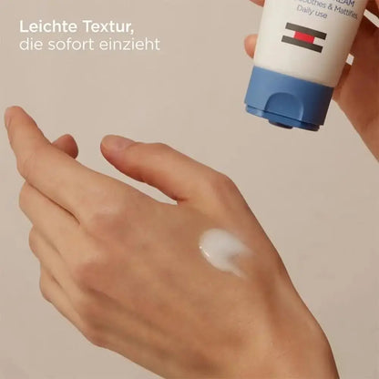 A hand holding a white and blue tube of body lotion above another hand showing a smear of the white cream. German text is visible on the top left.