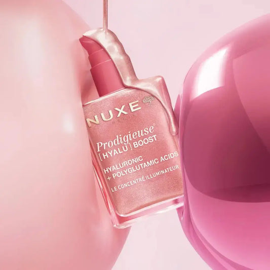 Nuxe Prodigieuse [Hyalýric] Boost hyaluronic and polyglutamic acid cream bottle in a pink shade, styled with glossy pink liquid and abstract pink spheres.