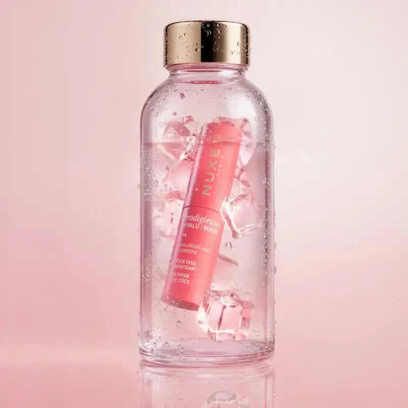 A clear glass bottle with a metallic gold cap, filled with ice and pink liquid, featuring a pink tube of NUXE Radiance Lip Balm floating inside. The bottle is centered against a soft pink background with a subtle reflection at the bottom.
