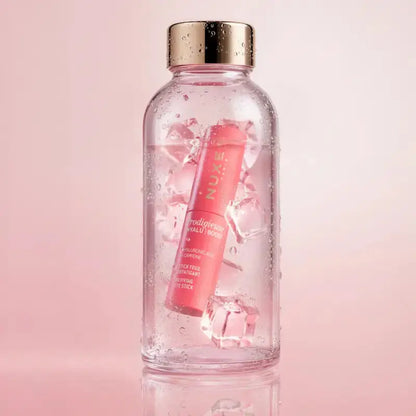 A clear glass bottle with a metallic gold cap, filled with ice and pink liquid, featuring a pink tube of NUXE Radiance Lip Balm floating inside. The bottle is centered against a soft pink background with a subtle reflection at the bottom.