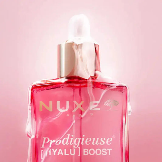 A close-up of a Nuxe Paris 'Prodigieuse [Hyalu] Boost' dropper bottle filled with pink liquid, set against a soft pink background.