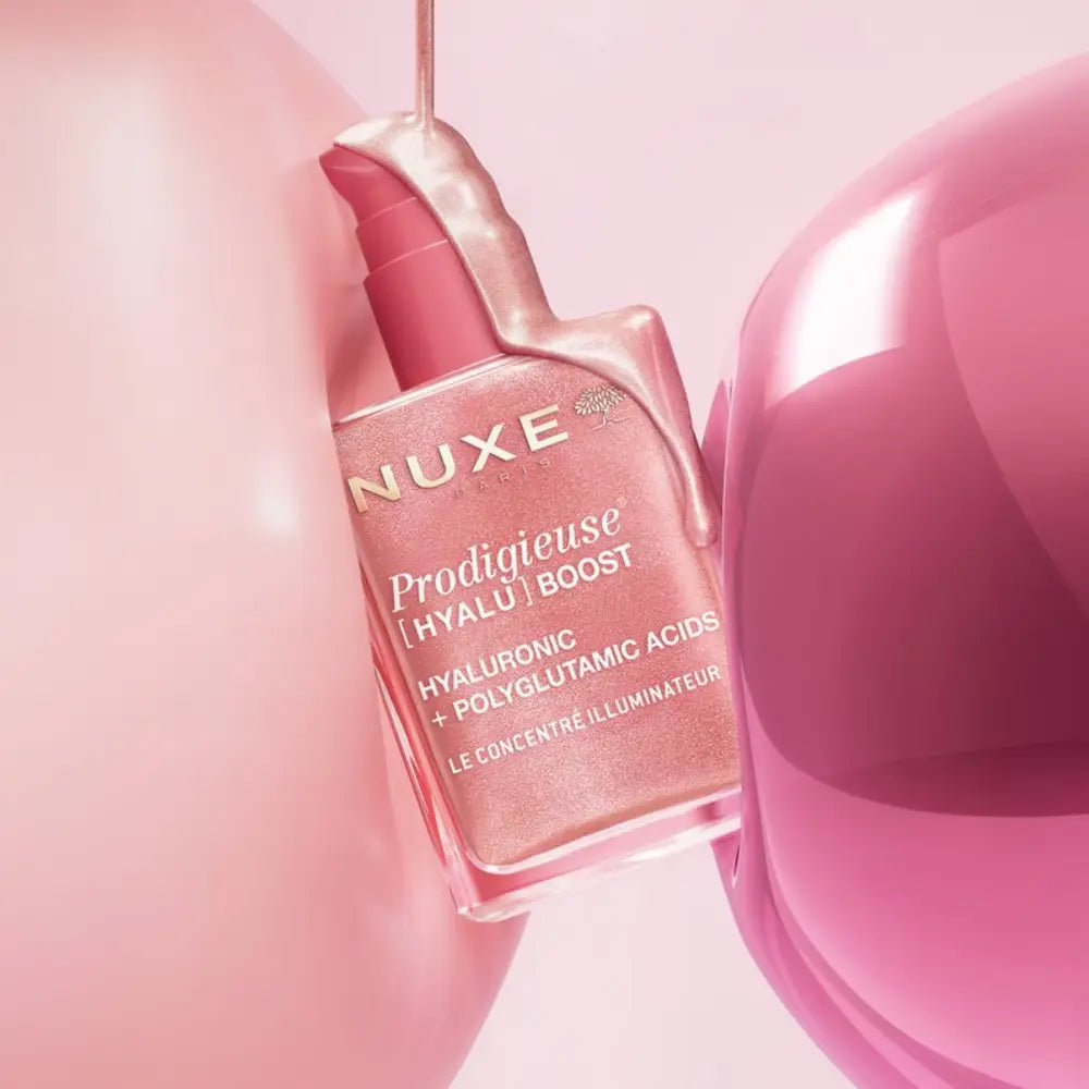 Nuxe Prodigieuse [Hyalýric] Boost hyaluronic and polyglutamic acid cream bottle in a pink shade, styled with glossy pink liquid and abstract pink spheres.