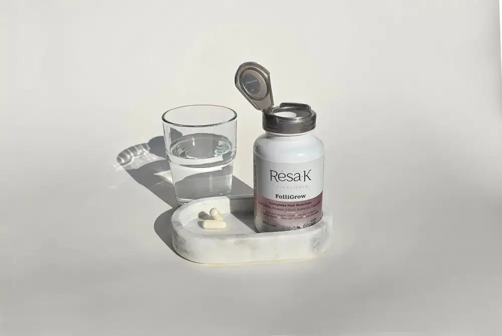 A bottle of ResaK Follitcrew dietary supplements with its cap open, shown next to a glass of water and a marble tray with a few capsules on a clean white background.
