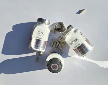 Two white supplement bottles labeled Resak Plus with capsules and one open cap arranged on a light gray surface with strong sunlight and shadows.