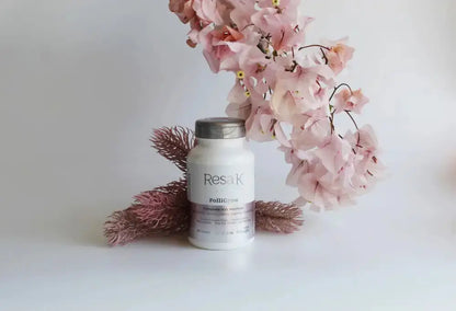 A white bottle of ResaK Polishing cream with a silver cap, styled on a white surface with pink flowers and dried brown grasses against a white background.