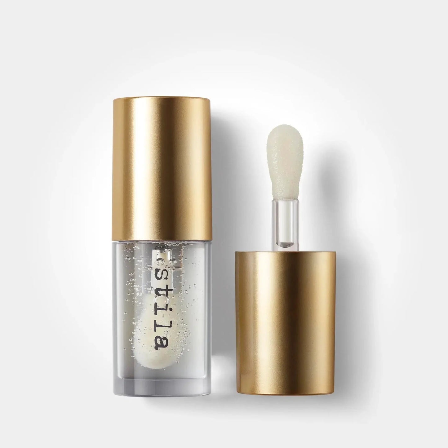 A clear cosmetic bottle with a metallic gold cap stands next to its applicator wand, which has a matching gold base and a soft, rounded brush tip. The bottle is transparent, showing a clear, bubbly white liquid inside, with the brand name 'bistilla' printed vertically in black on the lower half.