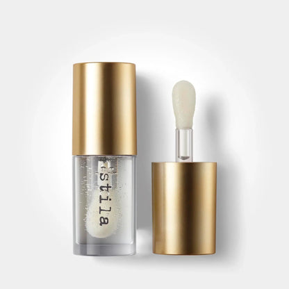 A clear cosmetic bottle with a metallic gold cap stands next to its applicator wand, which has a matching gold base and a soft, rounded brush tip. The bottle is transparent, showing a clear, bubbly white liquid inside, with the brand name 'bistilla' printed vertically in black on the lower half.