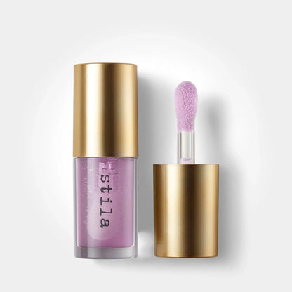 A clear bottle of lip gloss with a metallic gold cap, shown next to its applicator wand with a lavender-pink sponge tip. The brand name 'estila' is printed vertically on the bottle.