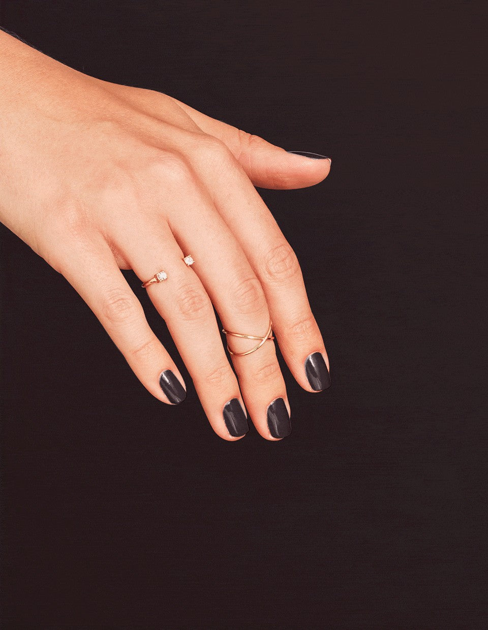 A close-up of a hand with dark manicured nails wearing three delicate gold rings against a solid black background.